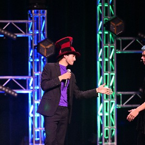 Actor with mic and black and red checkered hat stands on stage, holding a microphone in one hand and with his other outstretched, preparing for a handshake.