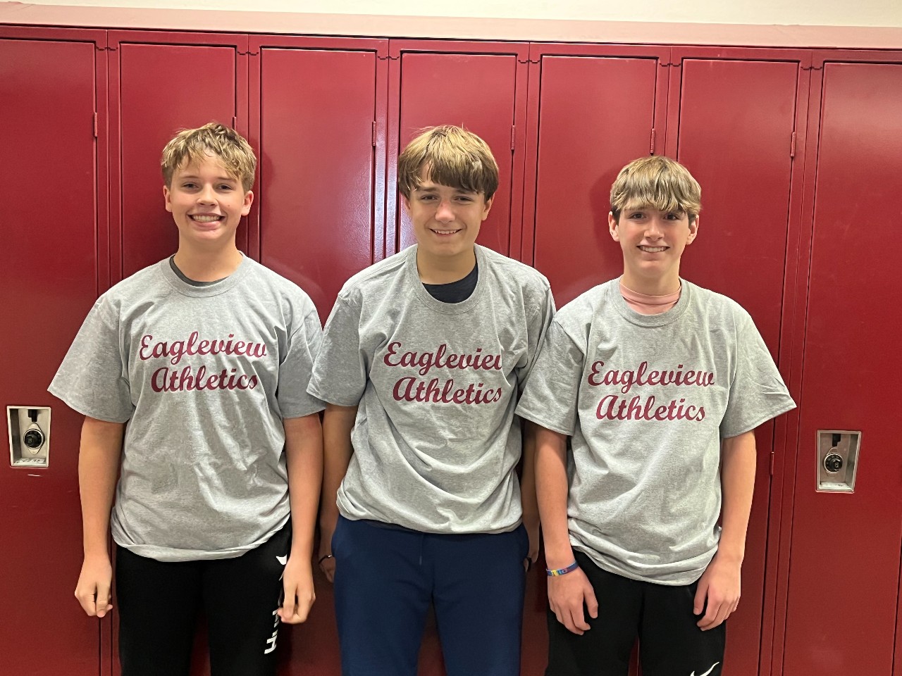 Three boys basketball players in their t-shirts stand in front of lockers.