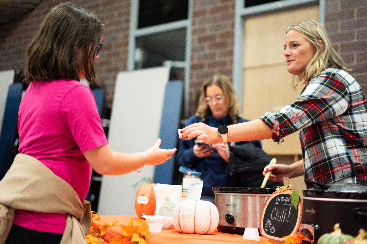 Liberty teacher Mrs. Dwinell hands a student a cup of chili from the crockpot during the Chili and Mac cook-off in Liberty's cafeteria. Assistant principal LiAnne Thiessen stands behind them.