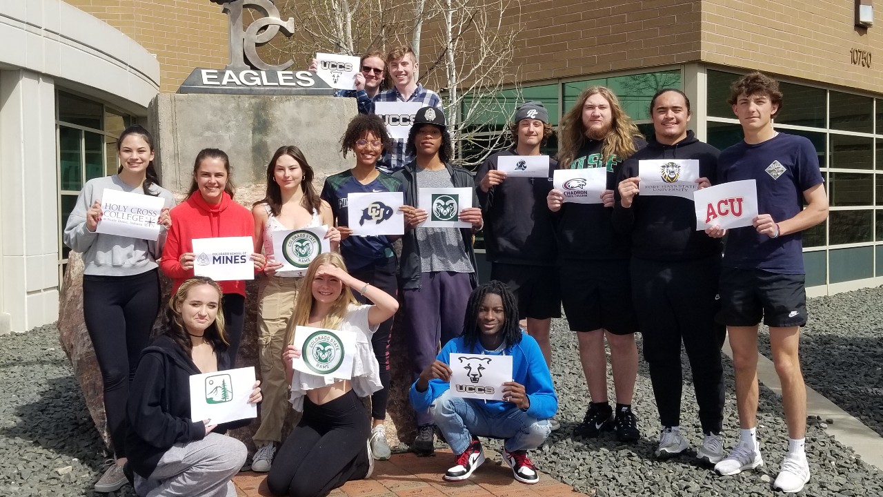 AVID students pose in front of school holding signs with the logo of the college they will attend in the fall, part 1