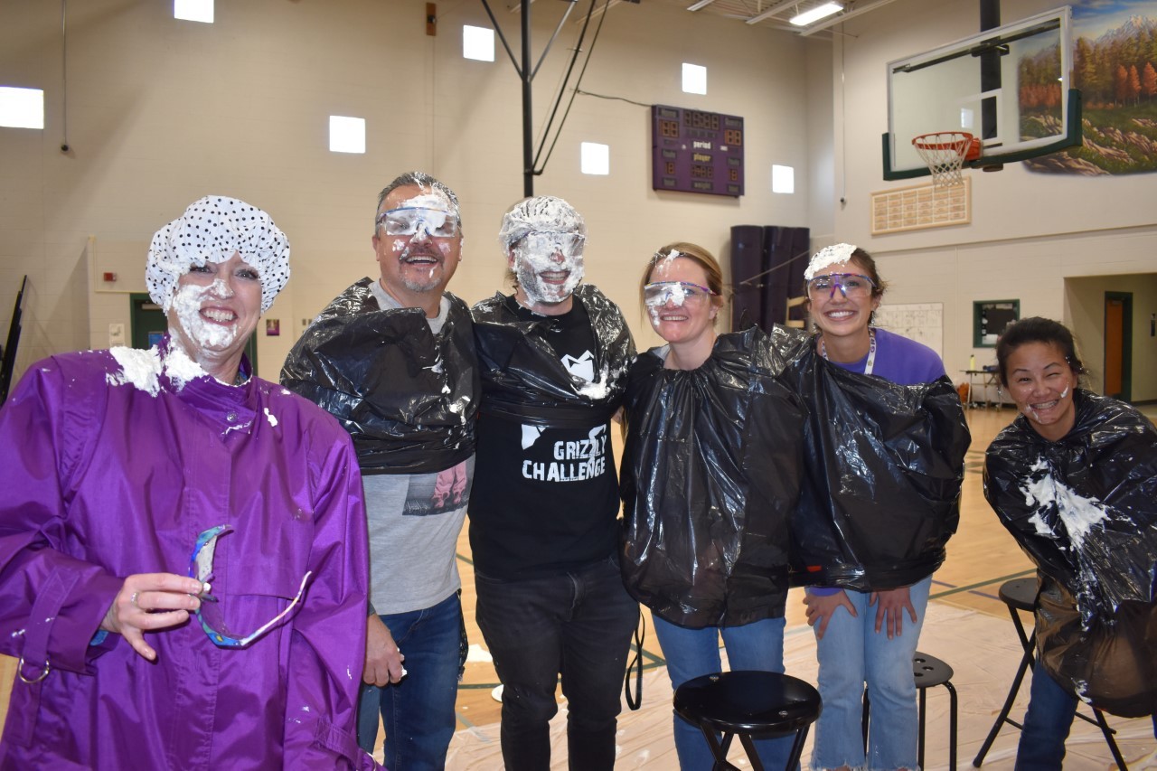 Six staff members standing in the gym wearing trash bags and they have pie all over their faces and hair