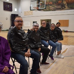 Six staff members sitting in the gym wearing trash bags and they are waiting to be pied in the face 