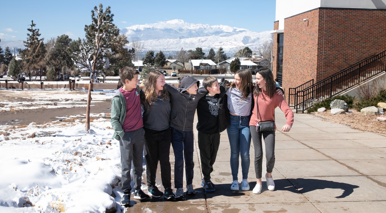 Six CMS students arm in arm near the school's entrance with a snow-covered Pikes Peak in the background.