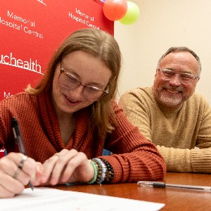 A father smiles at his daughter while she signs an agreement with UCHealth