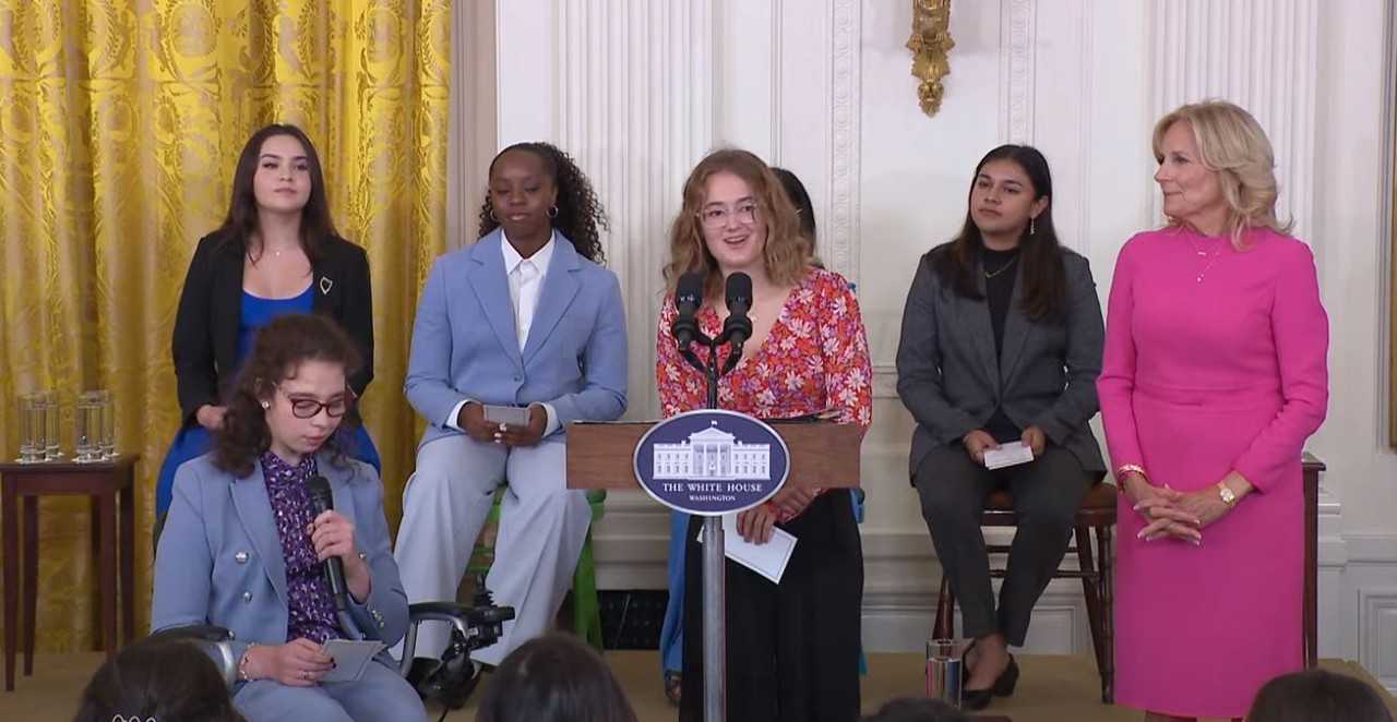 Liberty senior Avery Turner is shown in the White House press conference room speaking at the podium next to First Lady Jill Biden with other young women behind her.