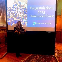 Hailey holds her scholarship certificate while standing in front of sign that says, "Congratulations 2023 Daniels Scholars!"