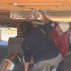 Two students work on the underside of a house floor to install HVAC components.