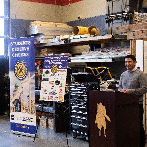 Liberty's construction teacher Jaime Hernandez stands behind a podium in Liberty's construction workshop with banners from company sponsors beside him.