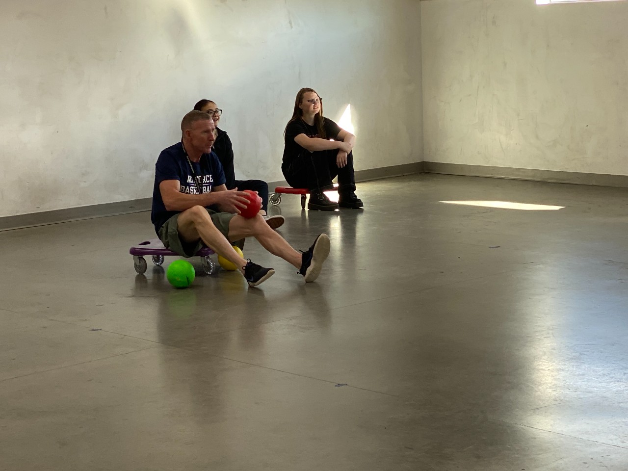 A teacher in an athletics room while sitting on a gym scooter and holding dodgeballs.