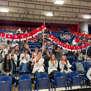 Liberty's robotics team sits in the gym stands at the BEST robotics competition with a "Lance-Up Robotics" banner above them.