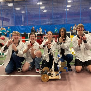Members of Liberty's Robotics team pose with the double L hand symbol in front of the competition field with their robot.
